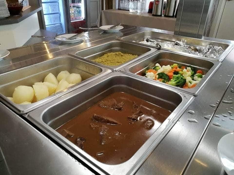 Stainless steel cafeteria buffet with trays of stew, boiled potatoes, mixed vegetables, and foil-covered food.