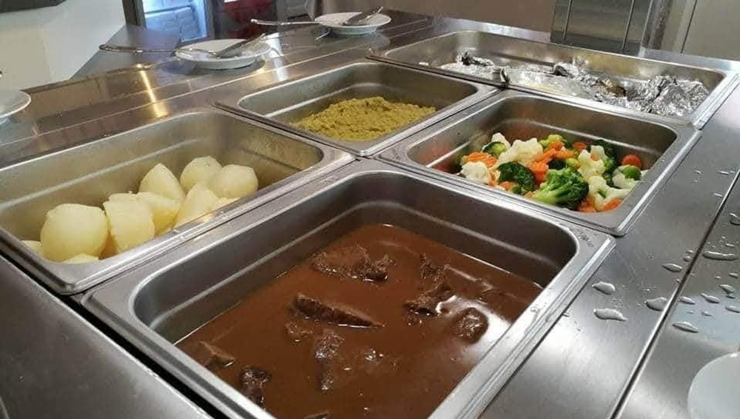 Stainless steel cafeteria buffet with trays of stew, boiled potatoes, mixed vegetables, and foil-covered food.