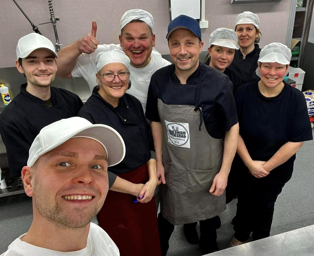 A group of employees in work uniforms standing together and smiling for a group photo in the kitchen.