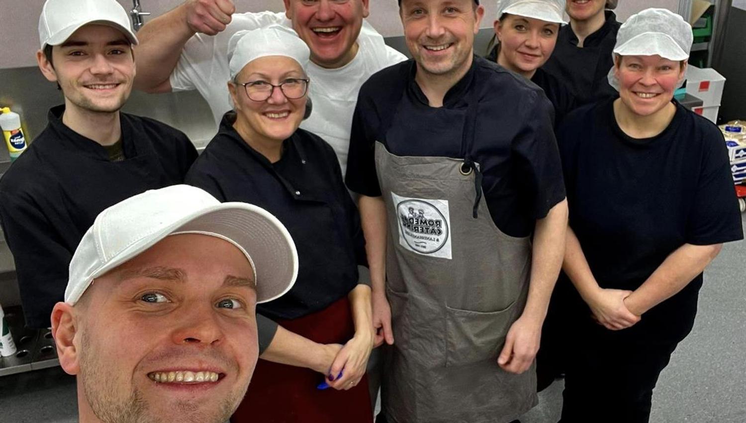 A group of employees in work uniforms standing together and smiling for a group photo in the kitchen.
