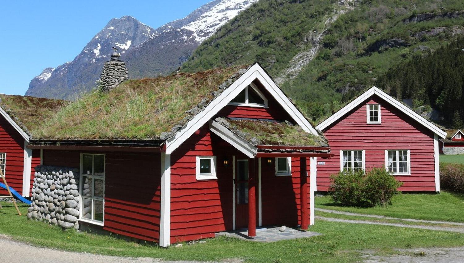 Two red wooden cabins with grass-covered roofs sit on green lawn, with forested and snowy mountains behind at Aabrekk Gard Trollbu in Oldedalen valley..