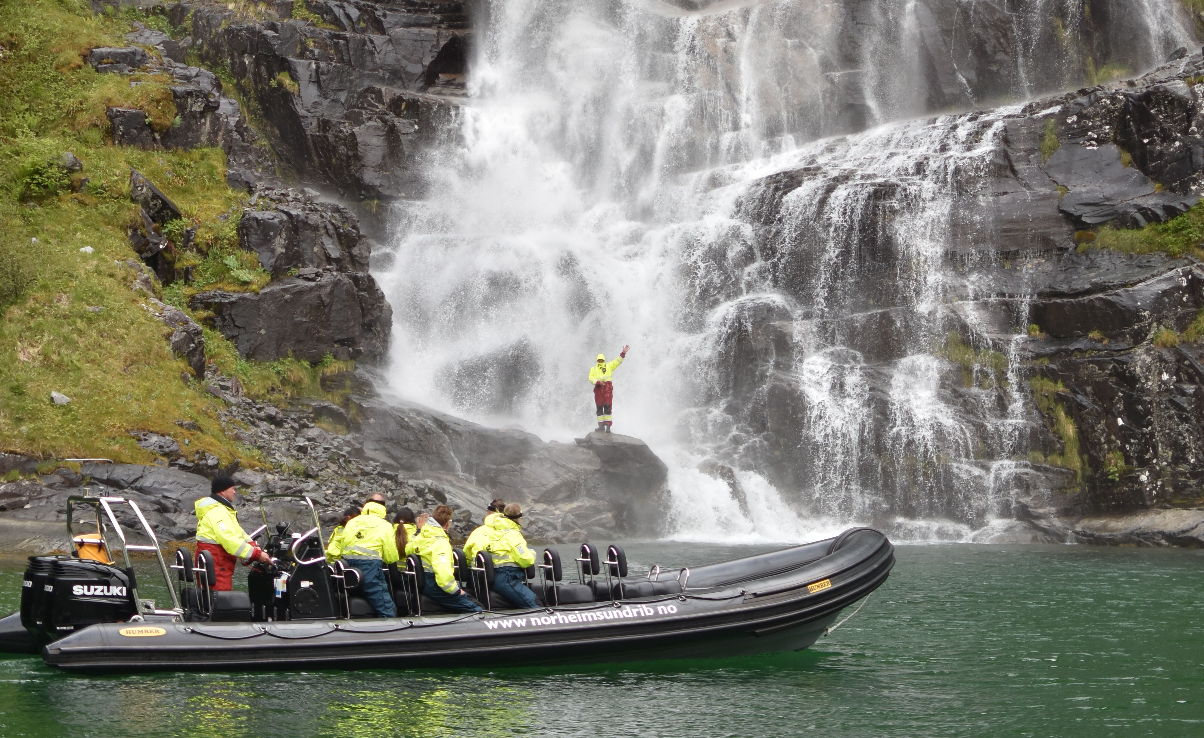 An adventurous participant stands on a rock near the waterfall while the rest of the group watches from the RIB boat.