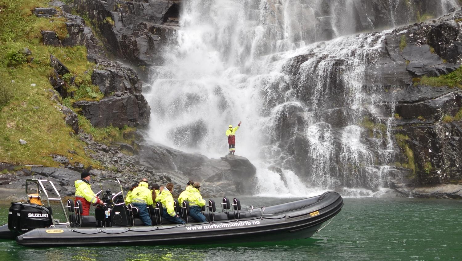 An adventurous participant stands on a rock near the waterfall while the rest of the group watches from the RIB boat.