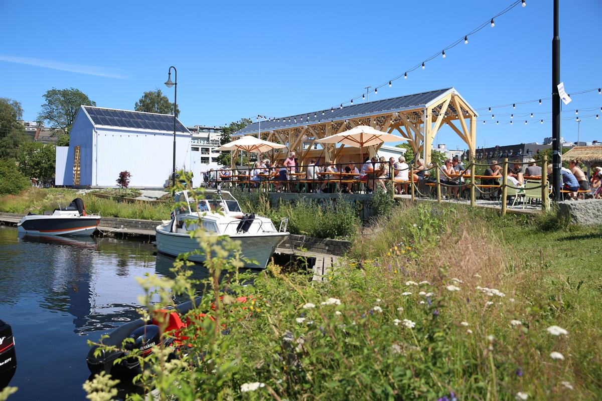 Uteservering og båter ved et idyllisk havneområde om sommeren.
