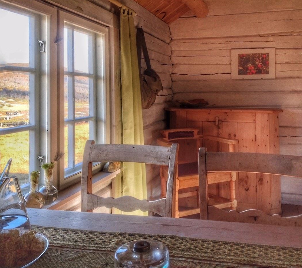 Indoor photo of a log‑style room with a wooden table and a window letting in warm daylight, surrounded by natural wood surfaces.