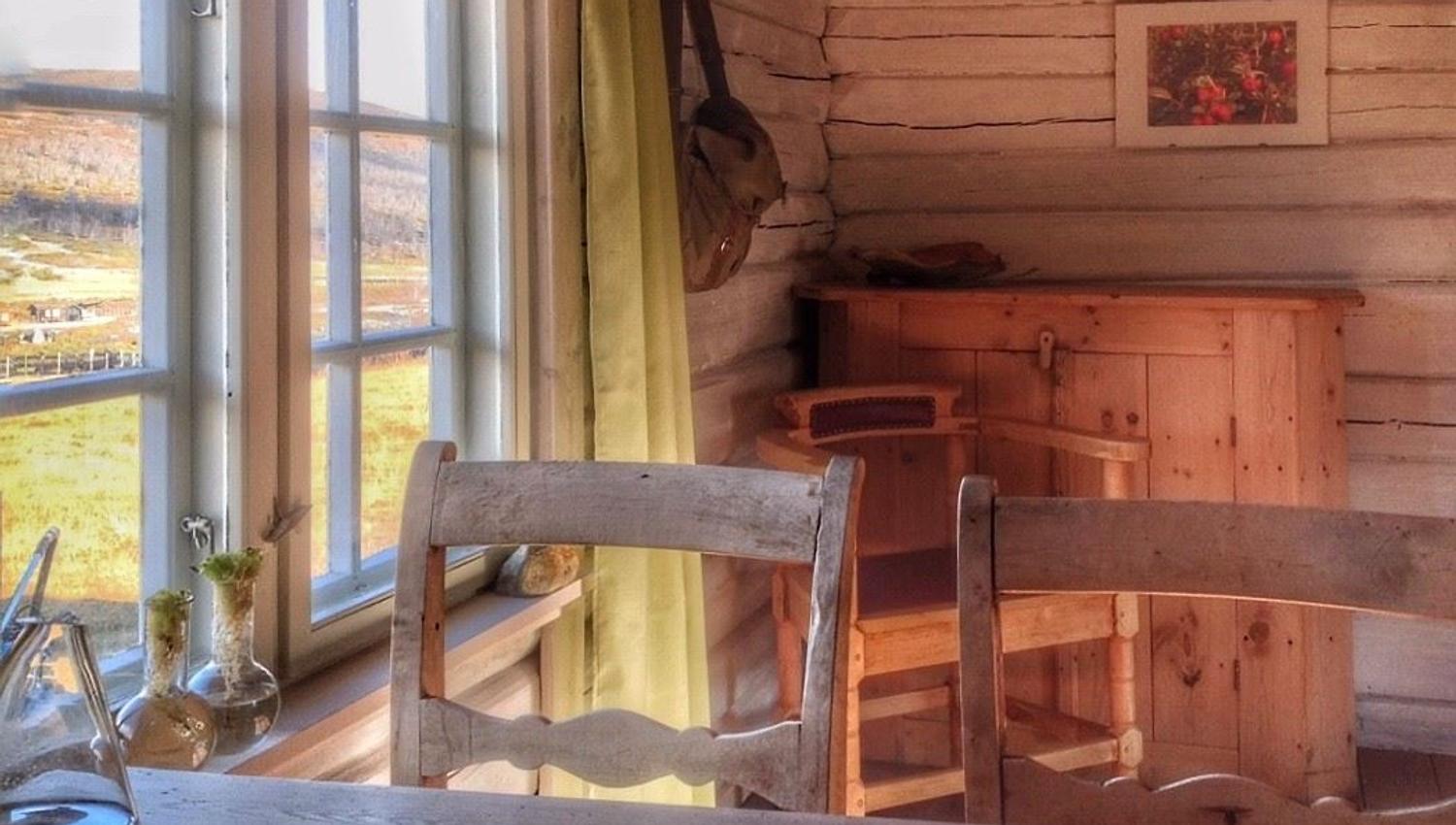 Indoor photo of a log‑style room with a wooden table and a window letting in warm daylight, surrounded by natural wood surfaces.