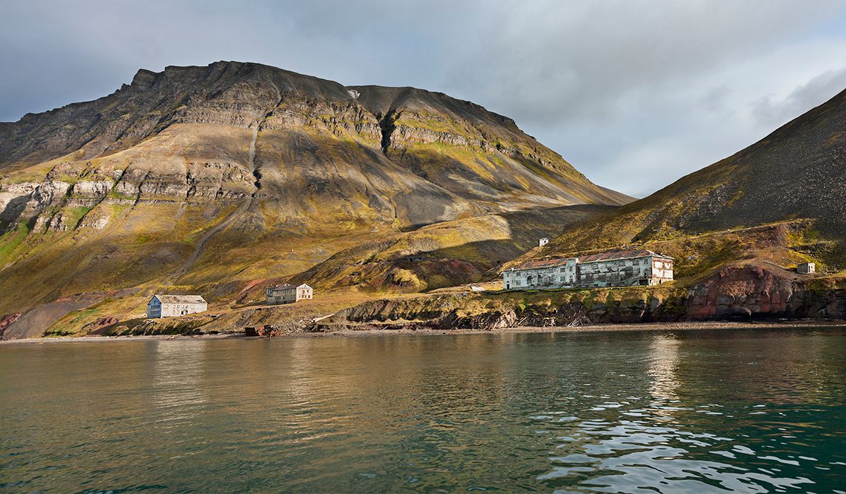 Forlatte bygninger i det tidligere gruvesamfunnet Grumant langs bredden av Isfjorden i forgrunnen, med høye fjell og en skyet himmel i bakgrunnen