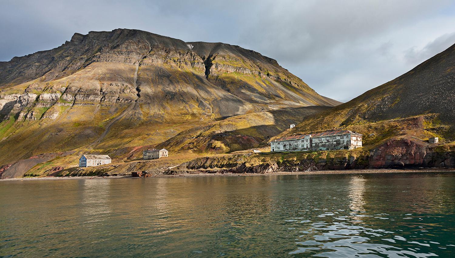 Forlatte bygninger i det tidligere gruvesamfunnet Grumant langs bredden av Isfjorden i forgrunnen, med høye fjell og en skyet himmel i bakgrunnen