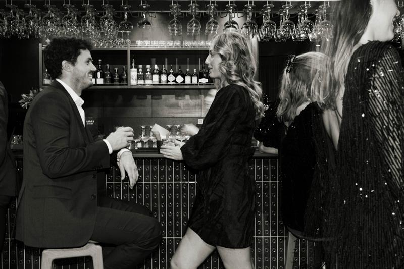 Mennesker kledd for fest samles ved en bar, noen i samtale og andre på vei forbi, i et elegant og livlig miljø.