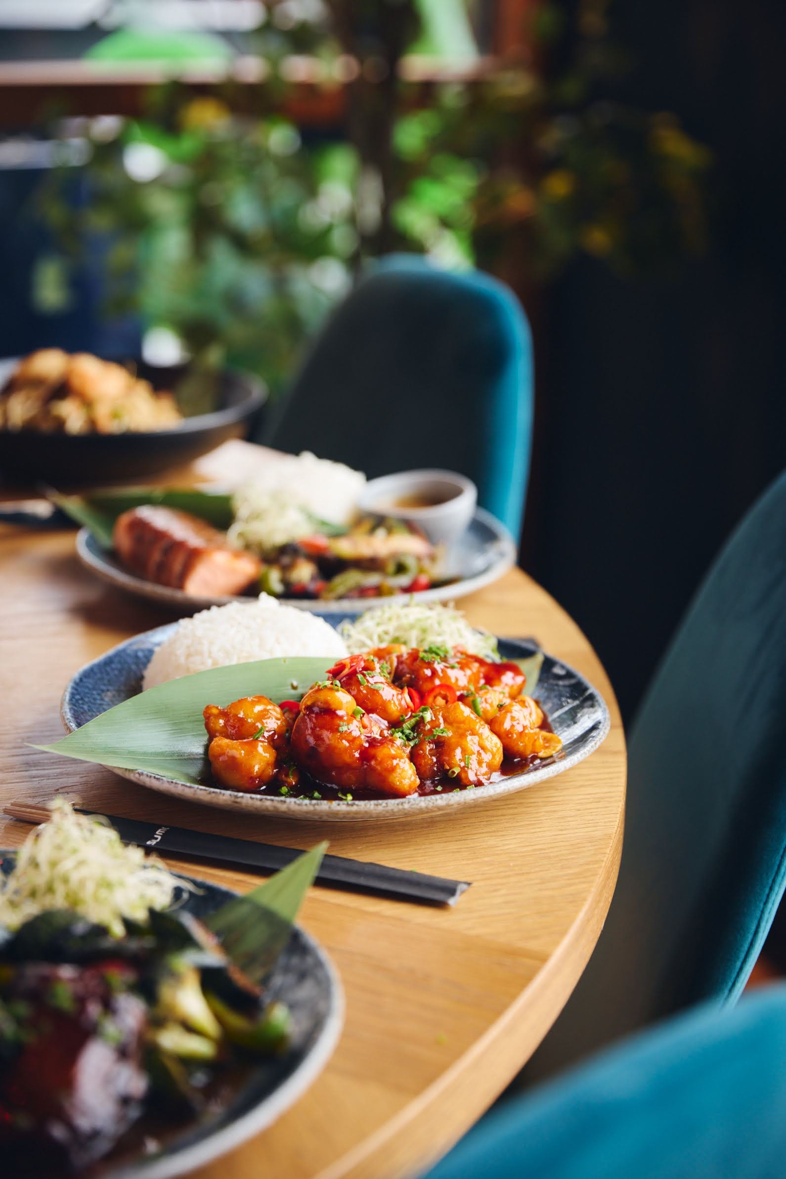 Nærbilde av restaurantbord med tallerkener med asiatisk mat, inkludert sursøt kylling med ris, laks og grønnsaker, omgitt av turkise stoler og grønt i bakgrunnen.