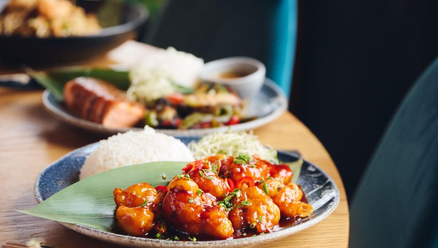 Nærbilde av restaurantbord med tallerkener med asiatisk mat, inkludert sursøt kylling med ris, laks og grønnsaker, omgitt av turkise stoler og grønt i bakgrunnen.