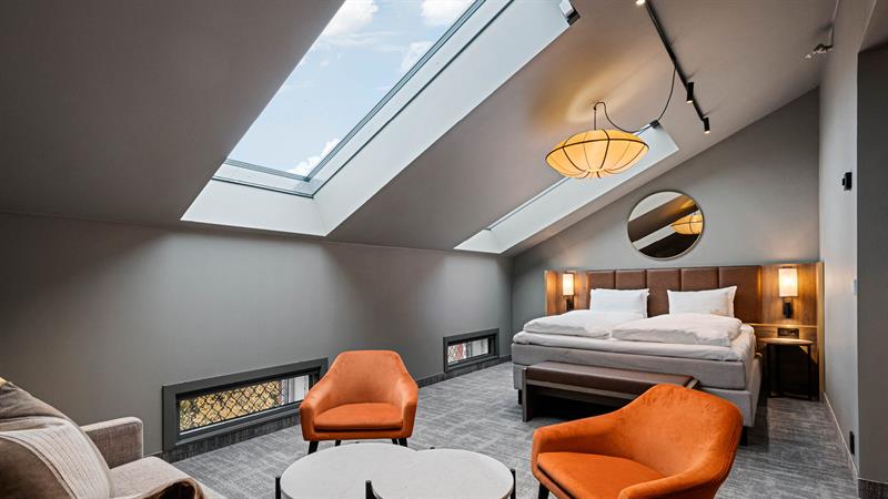 Loftsrom med skråtak, takvinuder oransje lenestoler, to små bord og beige sofa, og en stor dobbeltseng