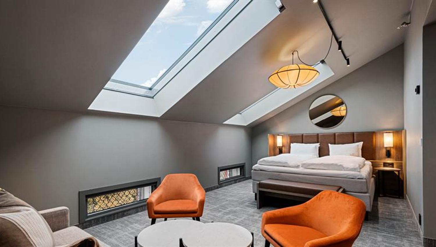 Loftsrom med skråtak, takvinuder oransje lenestoler, to små bord og beige sofa, og en stor dobbeltseng