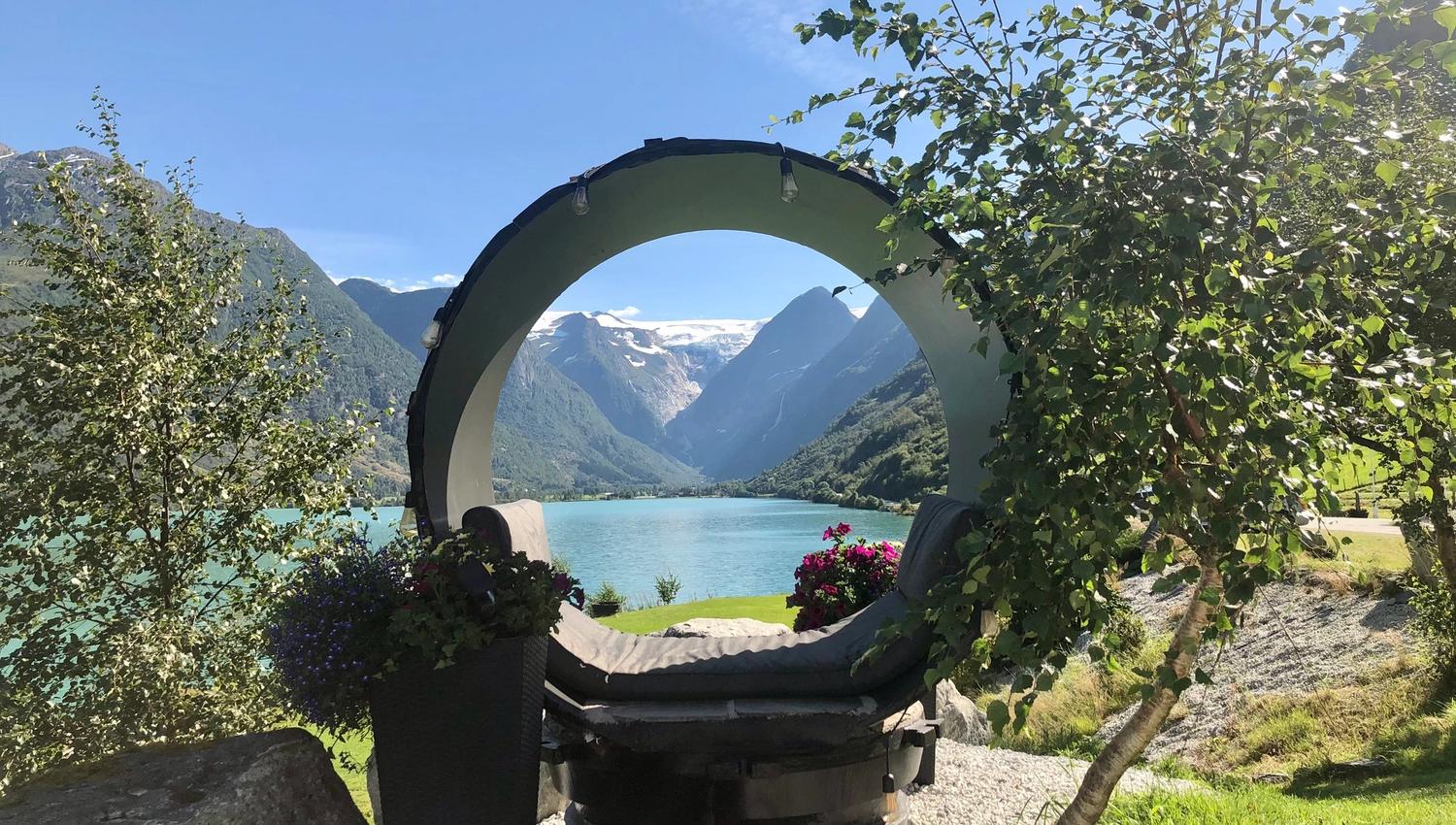 Circular outdoor lounge pod overlooks a turquoise lake and steep mountains, framed by trees under a clear blue sky..
