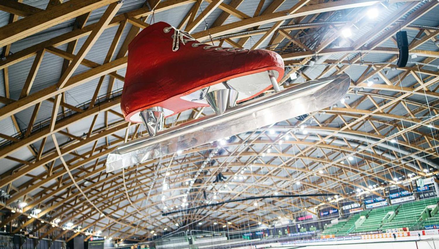 Indoor ice or multi-use arena with a wooden roof structure. A large red skate hangs decoratively above the rink.