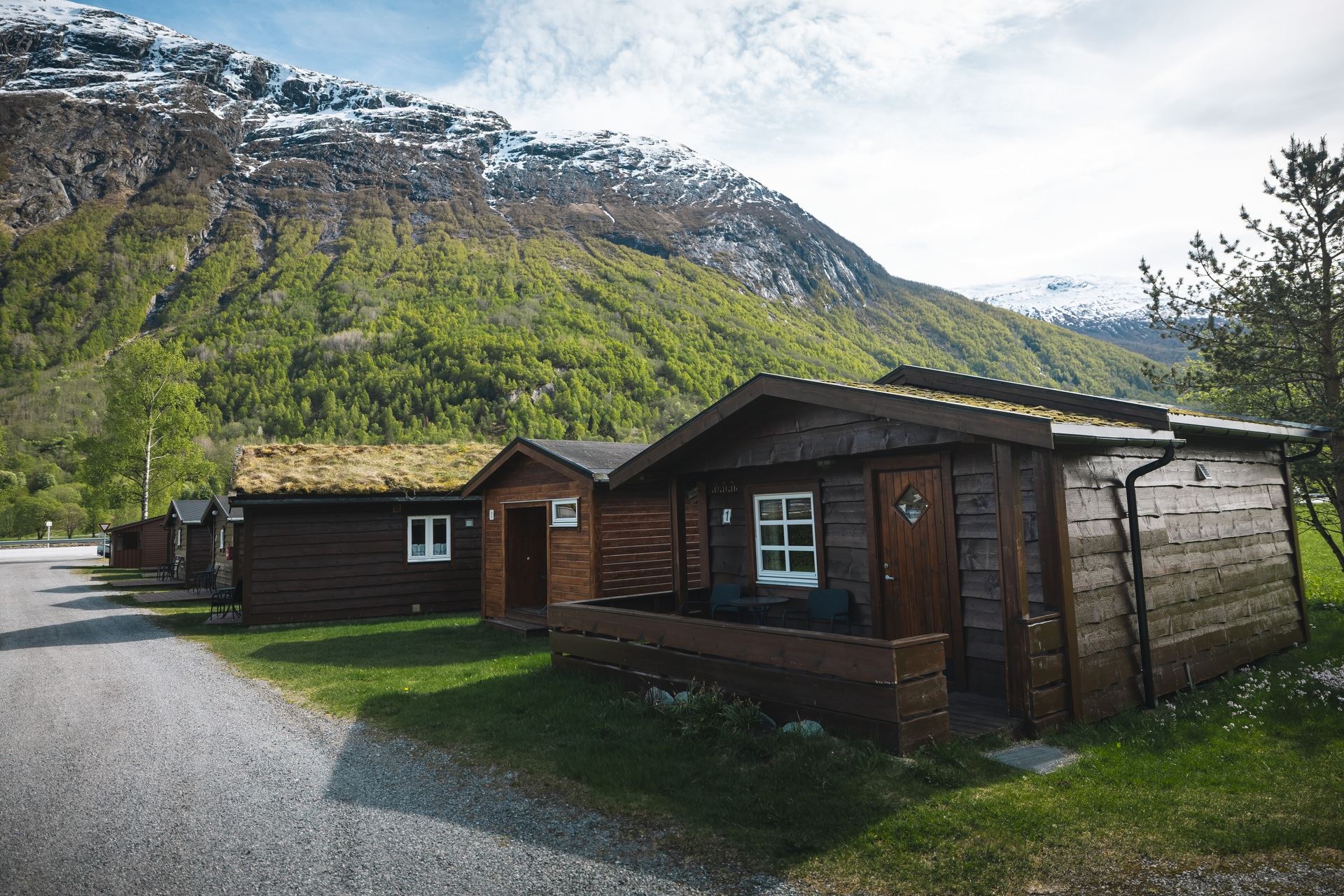 Wooden cabins with grass roofs beside a small road, set against green forested mountains with snowy peaks..