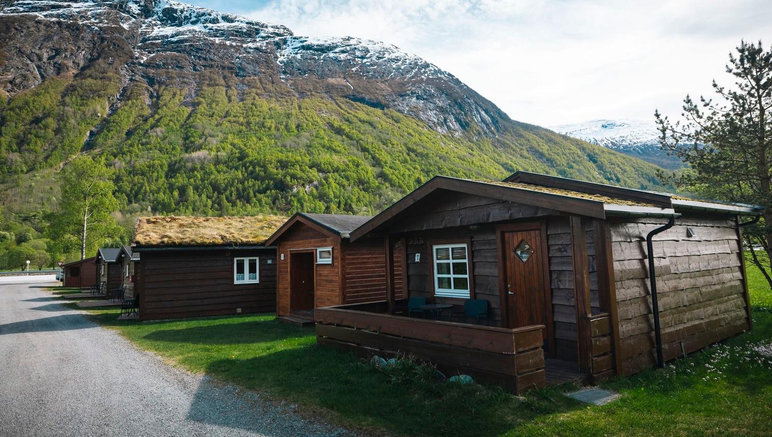 Wooden cabins with grass roofs beside a small road, set against green forested mountains with snowy peaks..