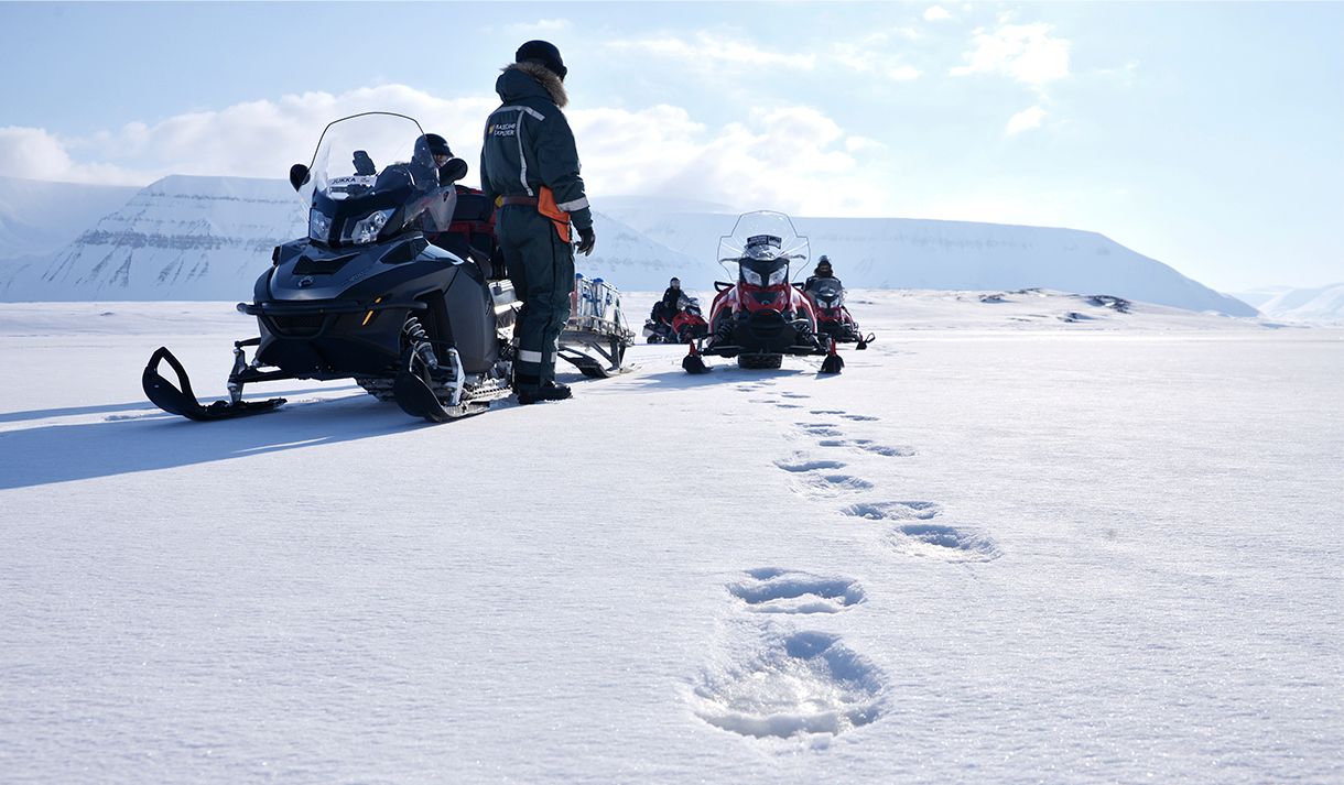 Polar bear tracks on a snowy plain in the foreground, with a guide and several snowmobiles in the background