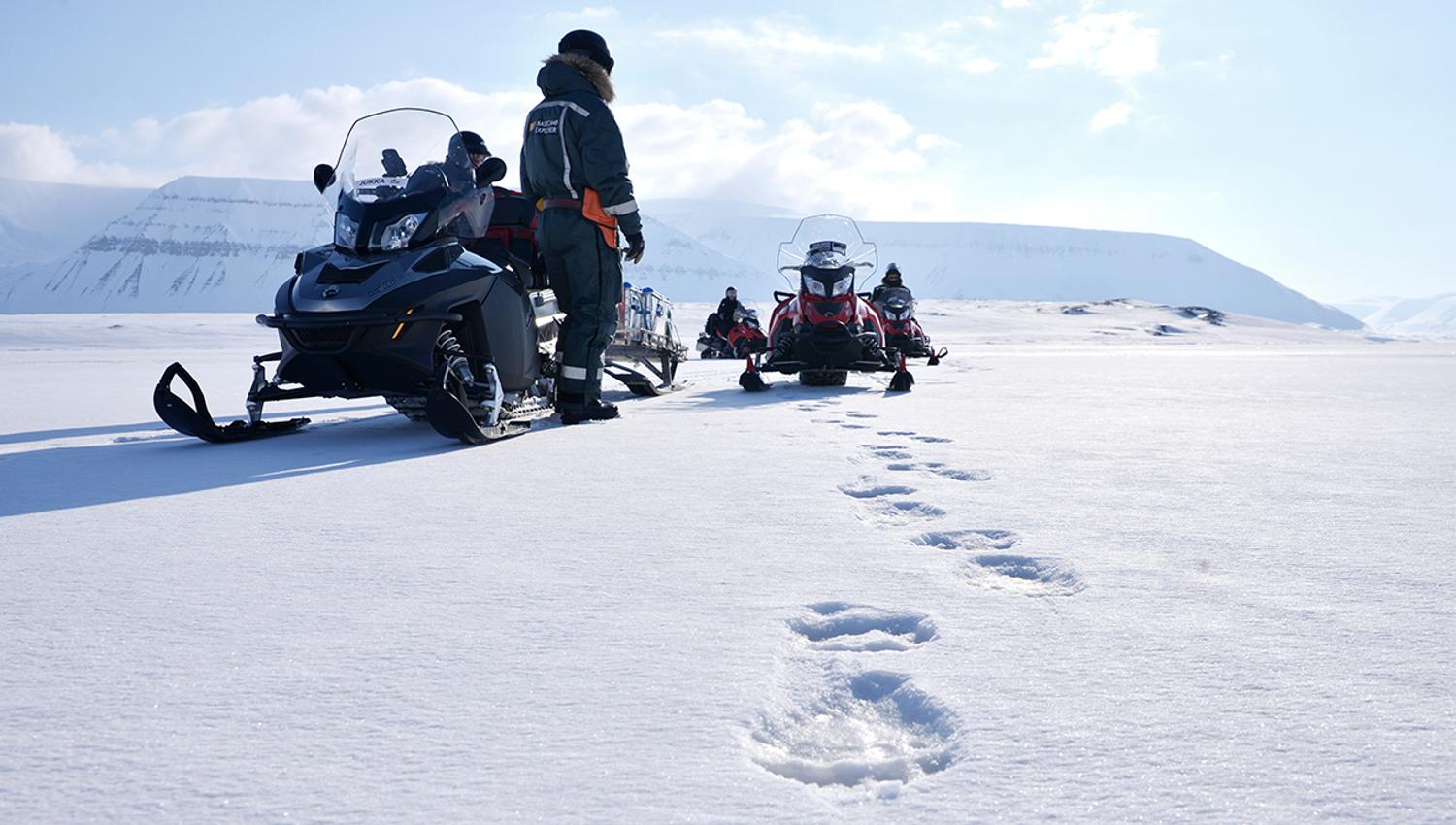 Polar bear tracks on a snowy plain in the foreground, with a guide and several snowmobiles in the background