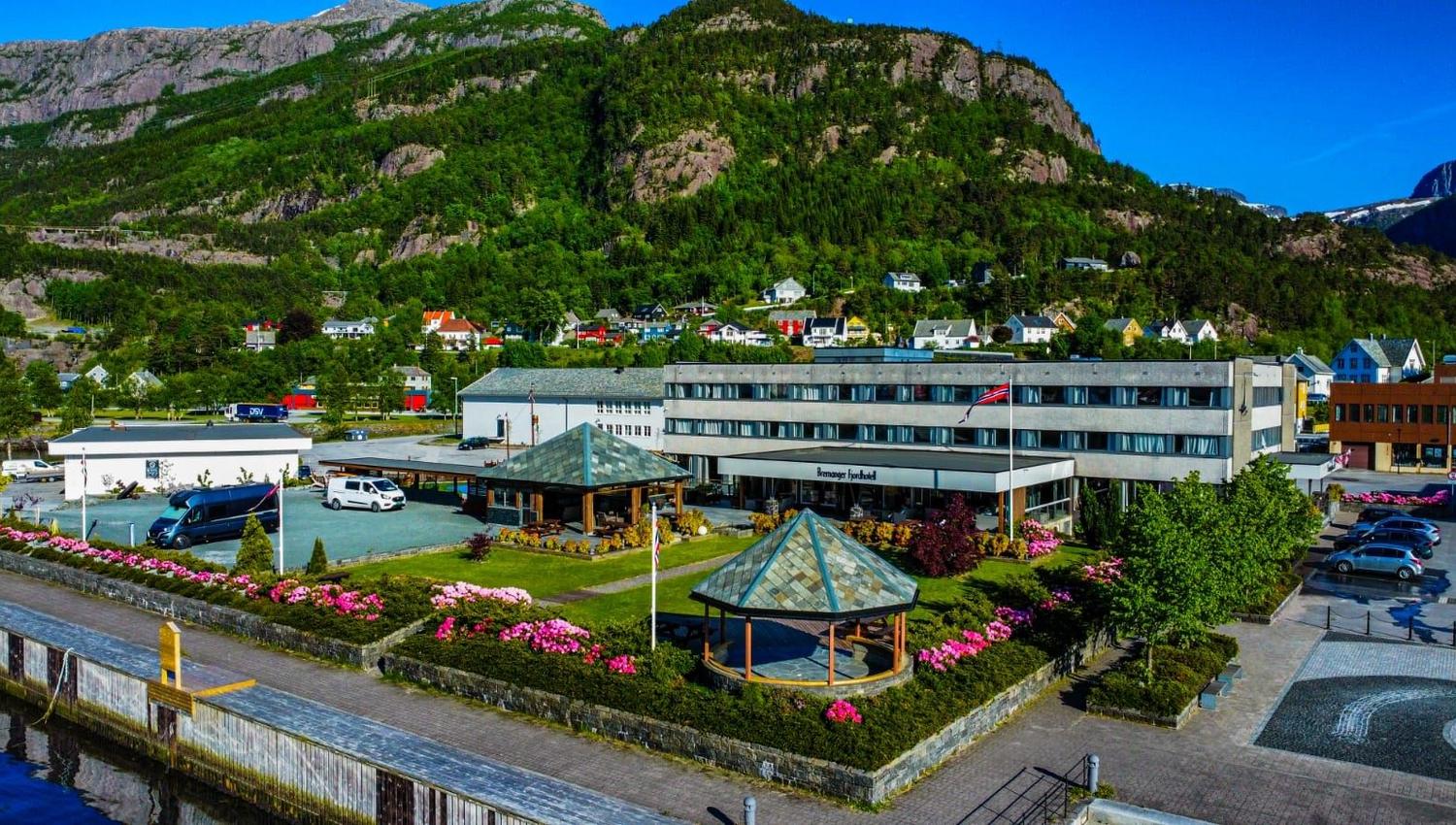 Bygningar med hage, flaggstong og parkering ved ei kai, med grøne fjell og bustadhus i bakgrunnen..