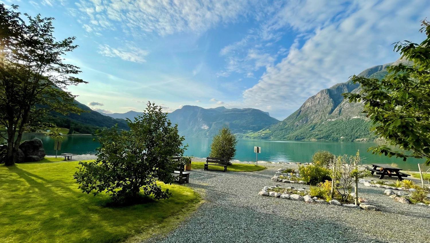 Grønt tun med grussti, benkar og tre ved ein roleg fjord, med bratte fjell og skya blå himmel..