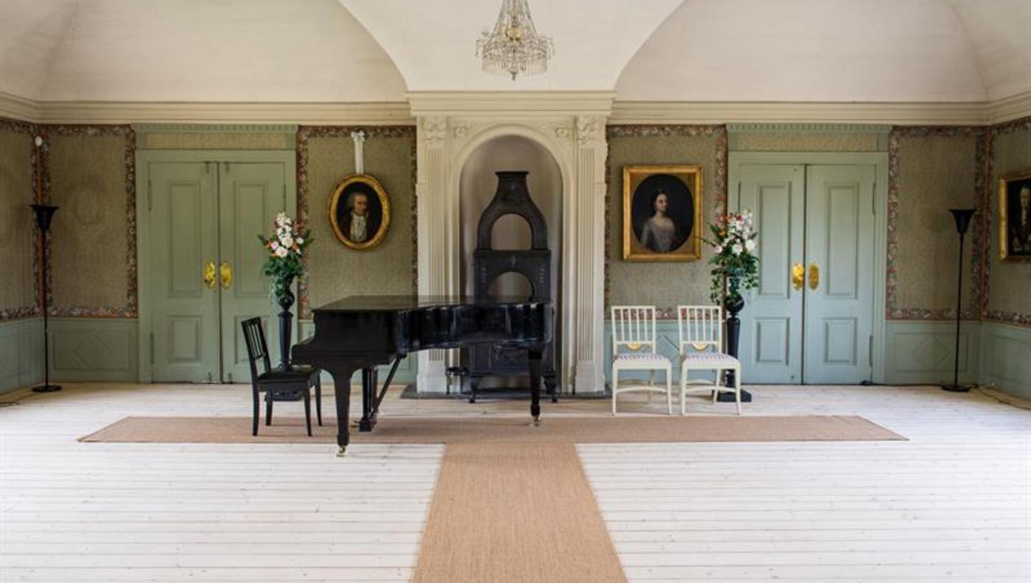 Historic hall featuring a grand piano, portraits and decorative details in a bright and symmetrical room.