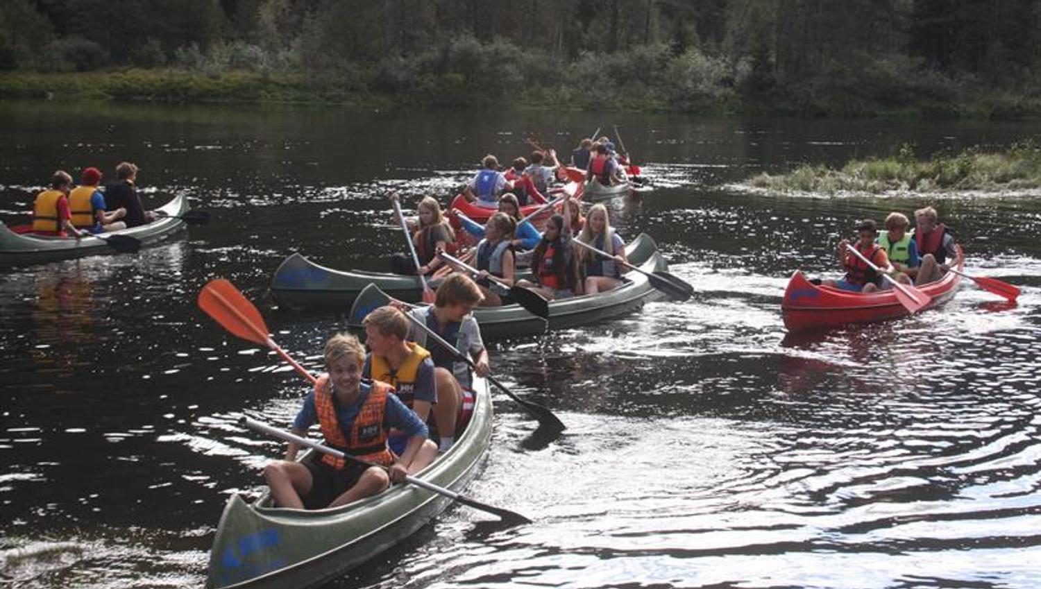 Eight canoes with three young people in each, paddling in the river