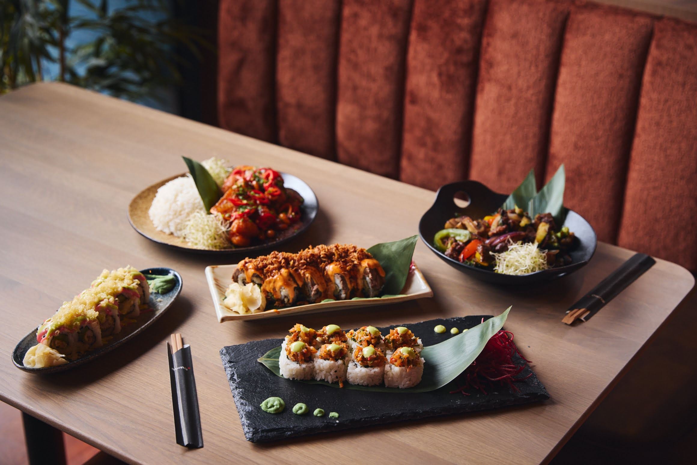 Trebord med fem asiatiske retter, inkludert sushi og garnityr, med spisepinner og en brun benk i bakgrunnen.