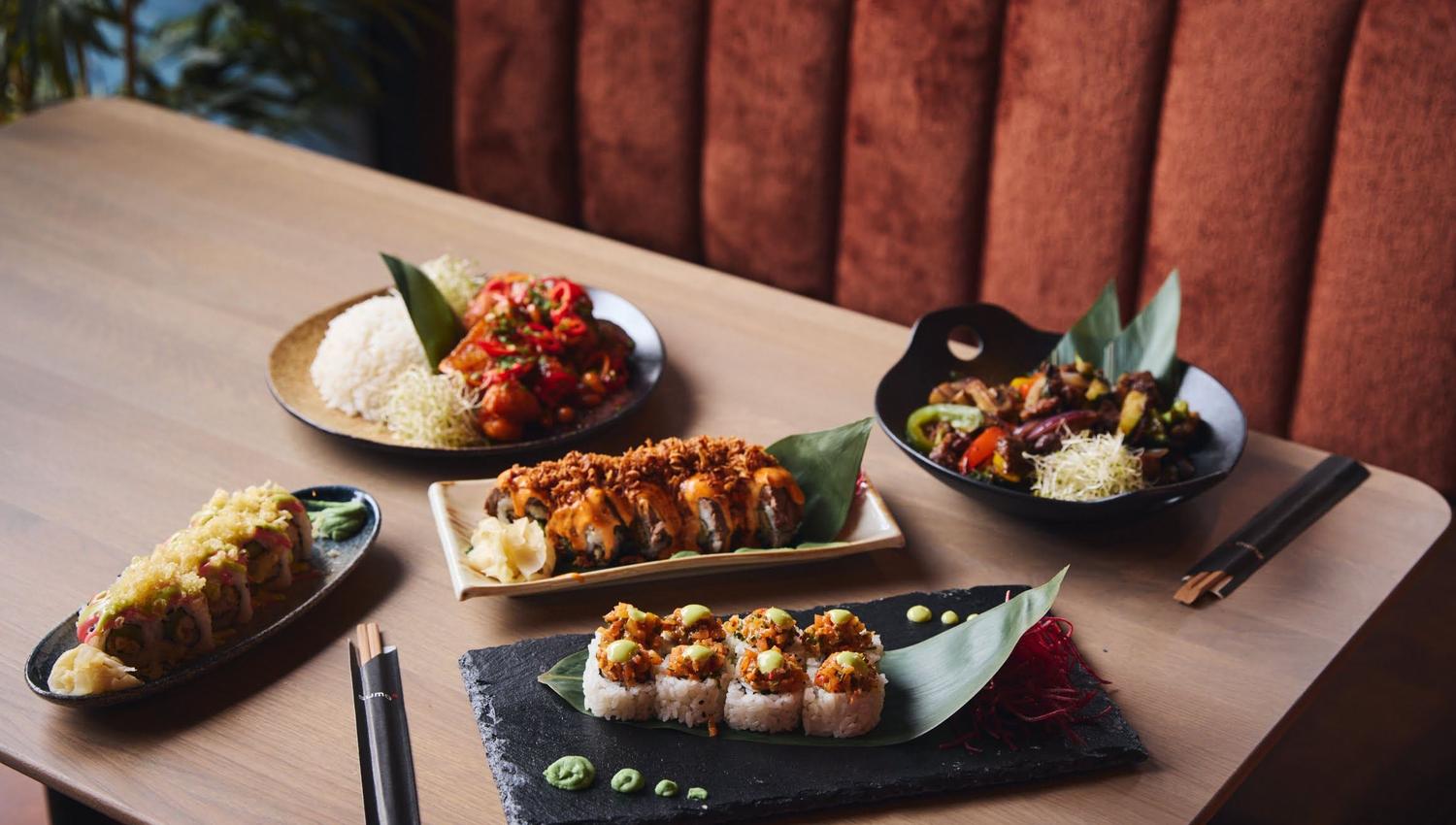 Trebord med fem asiatiske retter, inkludert sushi og garnityr, med spisepinner og en brun benk i bakgrunnen.