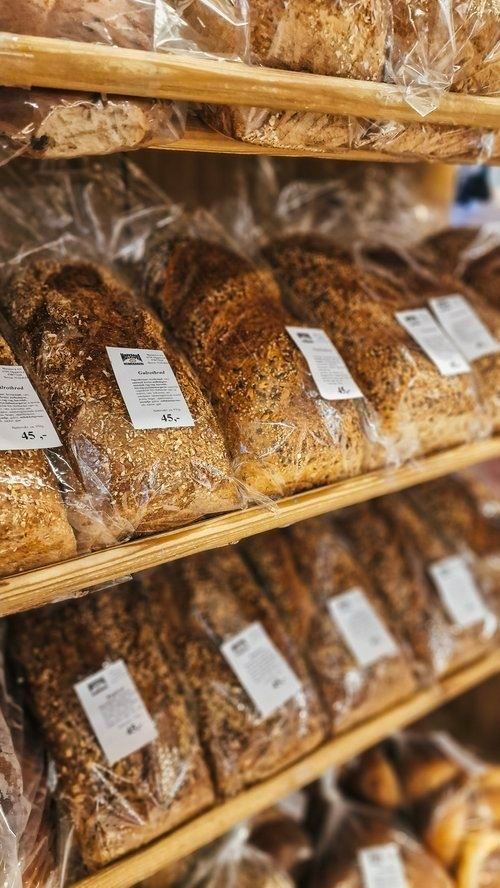 Rows of sliced and whole grain bread loaves in clear plastic packaging displayed on wooden bakery shelves with labels.