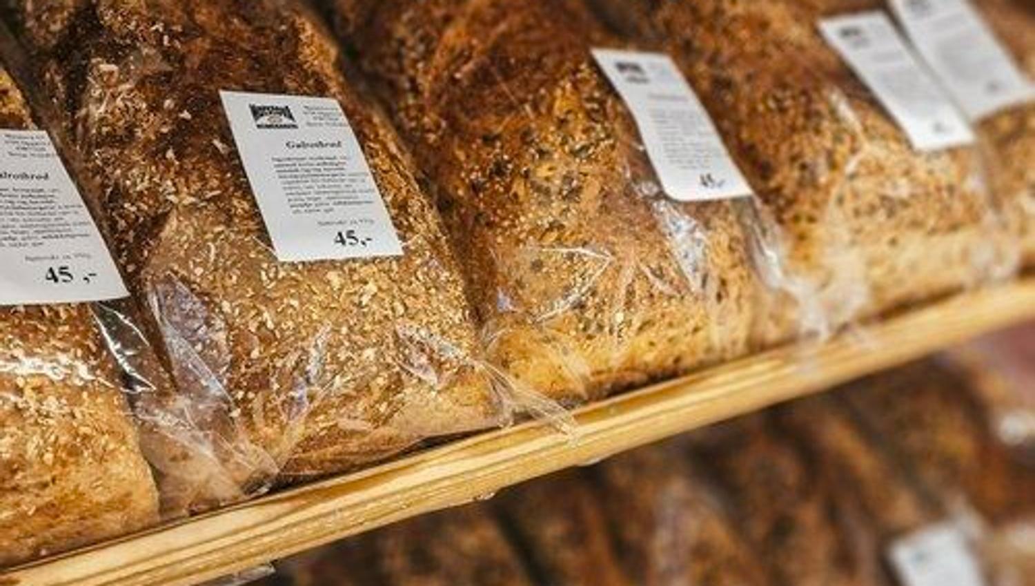 Rows of sliced and whole grain bread loaves in clear plastic packaging displayed on wooden bakery shelves with labels.