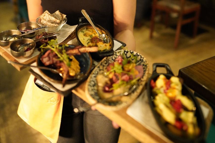 Brett med flere småretter, inkludert flatbrød, suppe, grillmat, salat og pastarett, i en innendørs restaurantsetting.