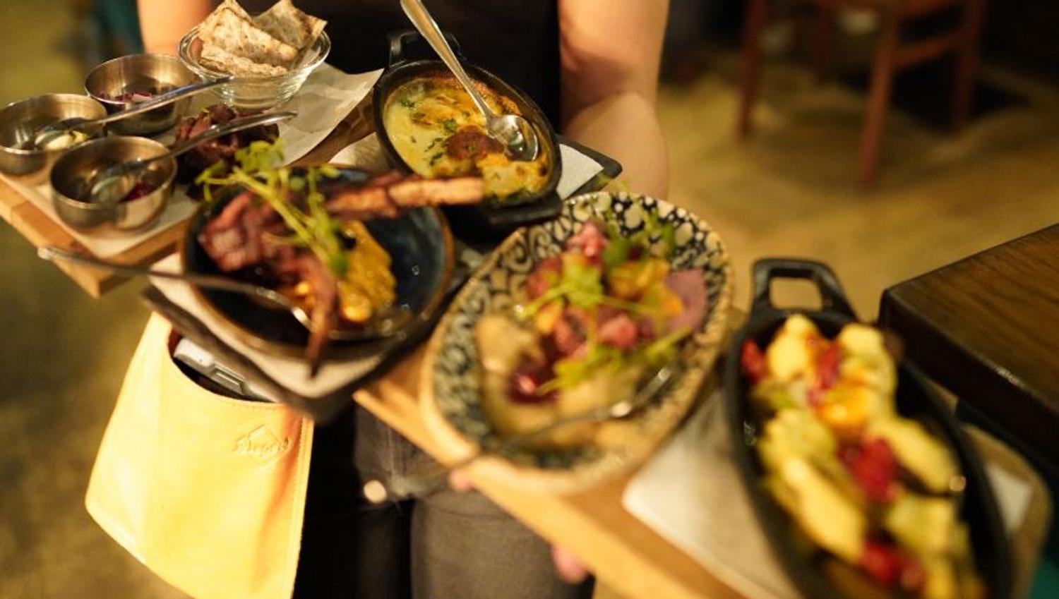 Brett med flere småretter, inkludert flatbrød, suppe, grillmat, salat og pastarett, i en innendørs restaurantsetting.