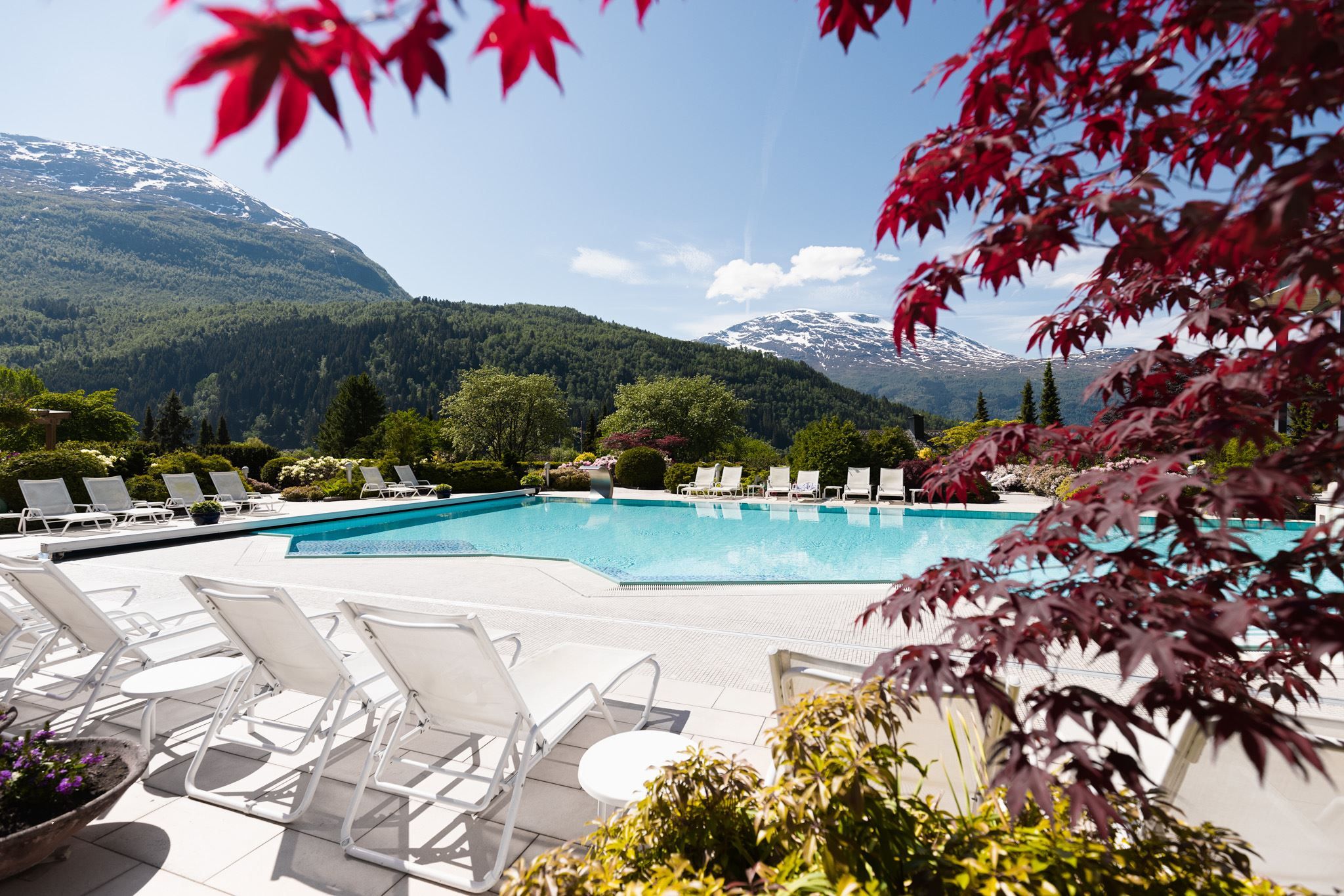 Outdoor swimming pool with white lounge chairs, gardens, and forested mountains with snow patches under a clear sky in Loen..