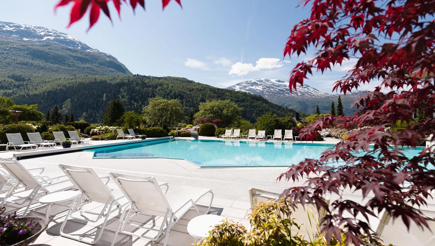 Outdoor swimming pool with white lounge chairs, gardens, and forested mountains with snow patches under a clear sky in Loen..