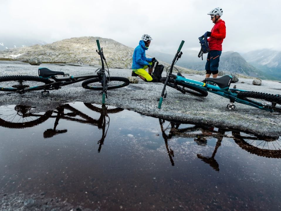 Two mountain bikers on a rocky mountain plateau by a pond, with bikes on the ground and misty mountains in the background.