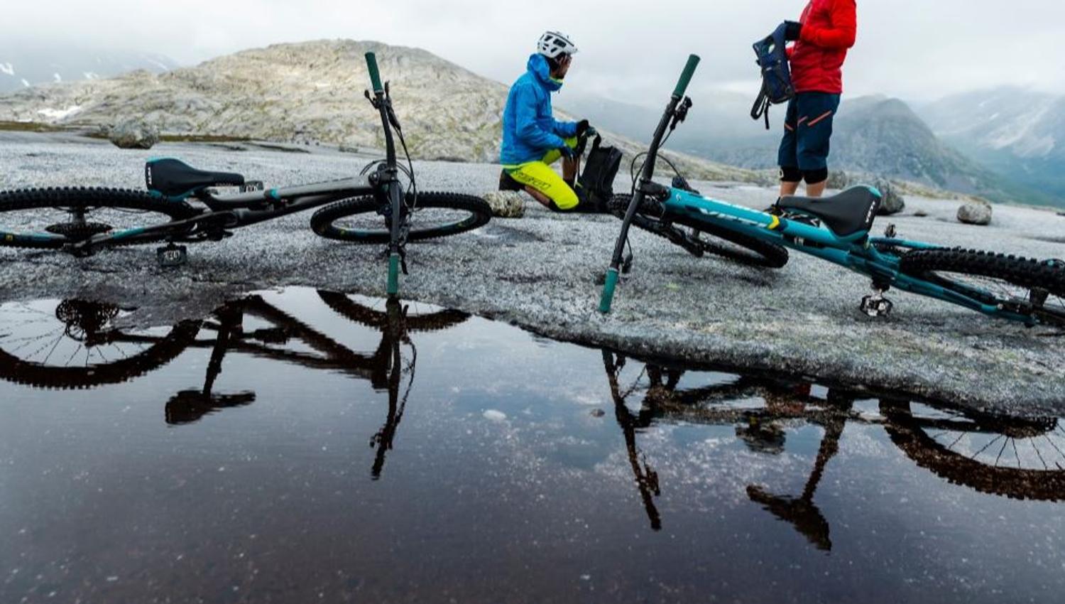 Two mountain bikers on a rocky mountain plateau by a pond, with bikes on the ground and misty mountains in the background.