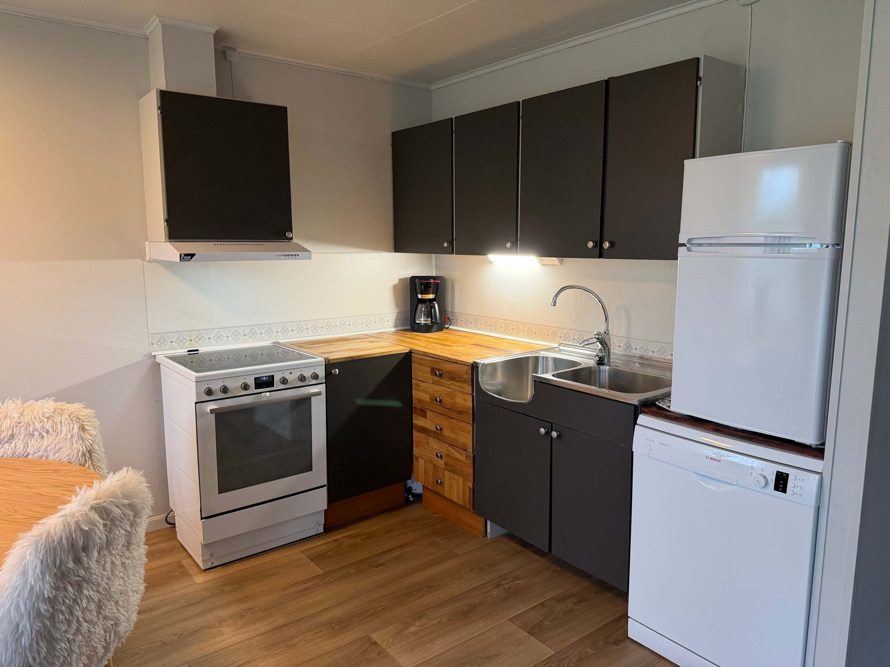 Small kitchen with black cabinets, wood countertop, stove, sink, coffee maker, fridge, and dishwasher on wood floor.