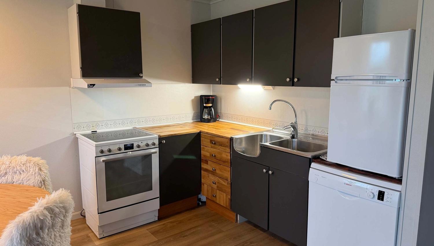 Small kitchen with black cabinets, wood countertop, stove, sink, coffee maker, fridge, and dishwasher on wood floor.