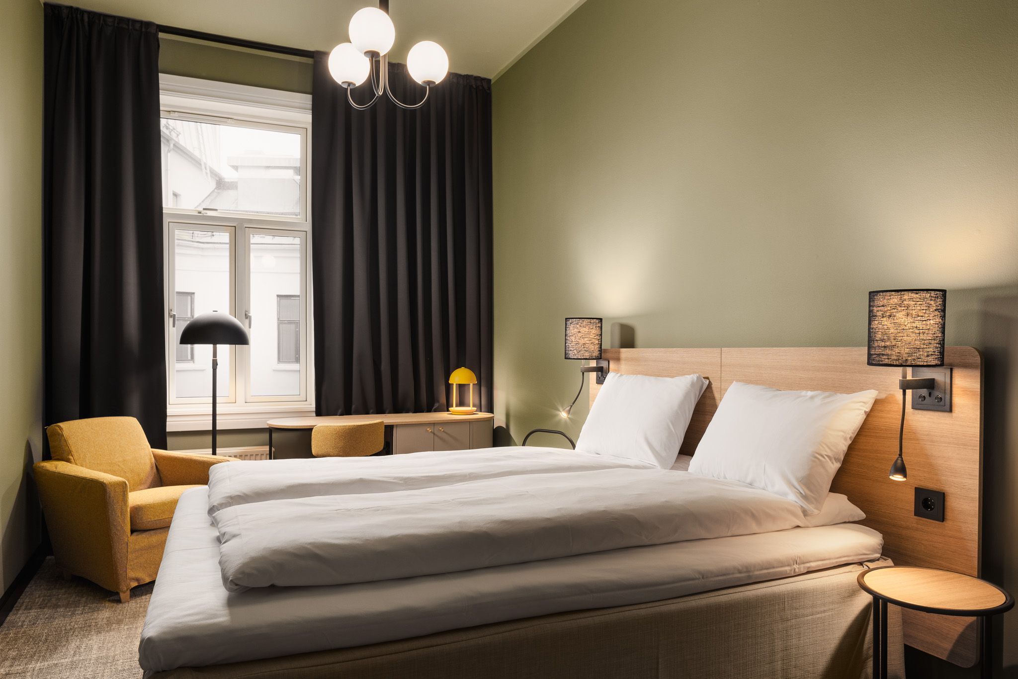 A green-painted hotel room with a double bed, a yellow armchair, a writing desk, and black curtains