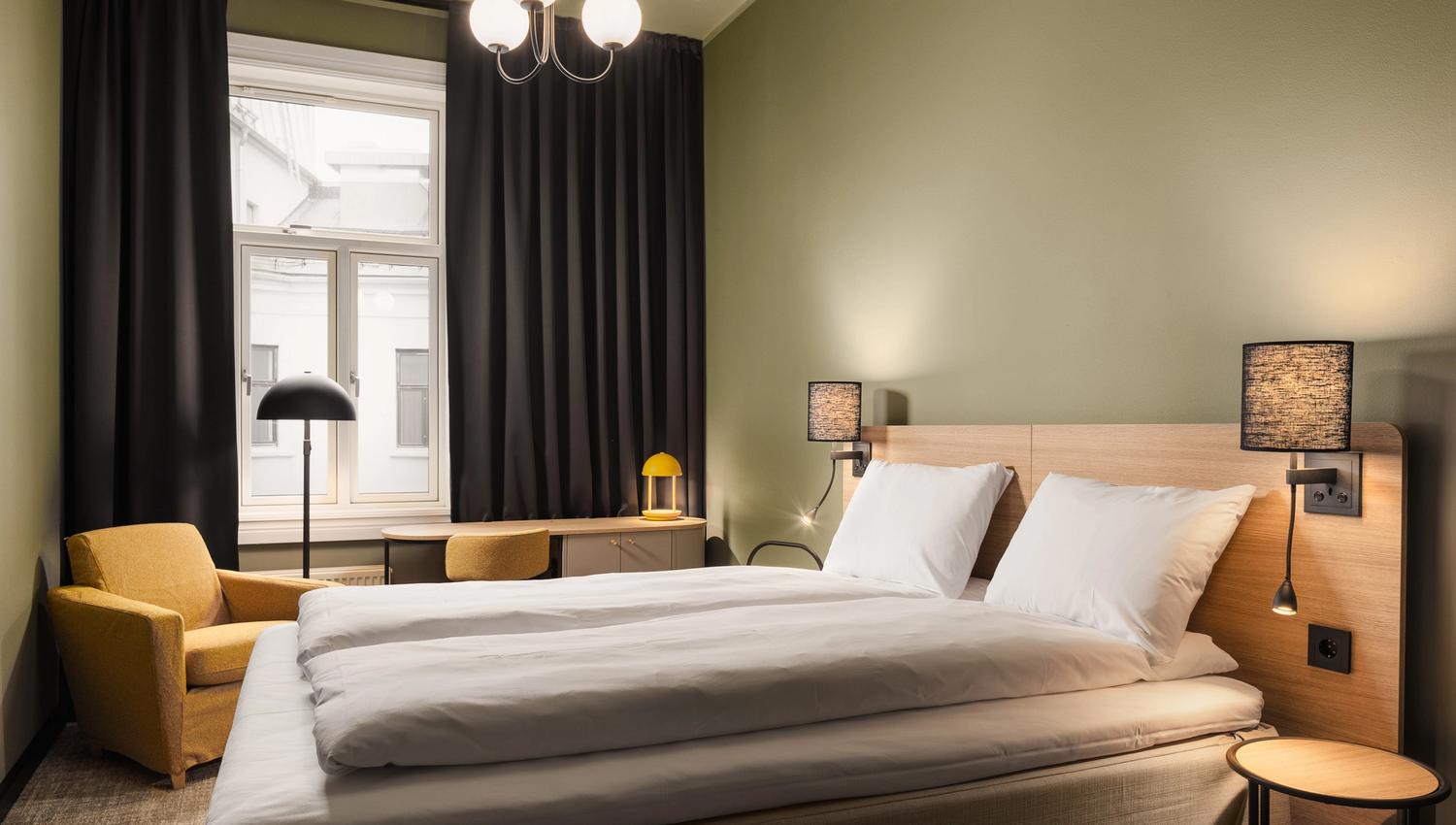 A green-painted hotel room with a double bed, a yellow armchair, a writing desk, and black curtains