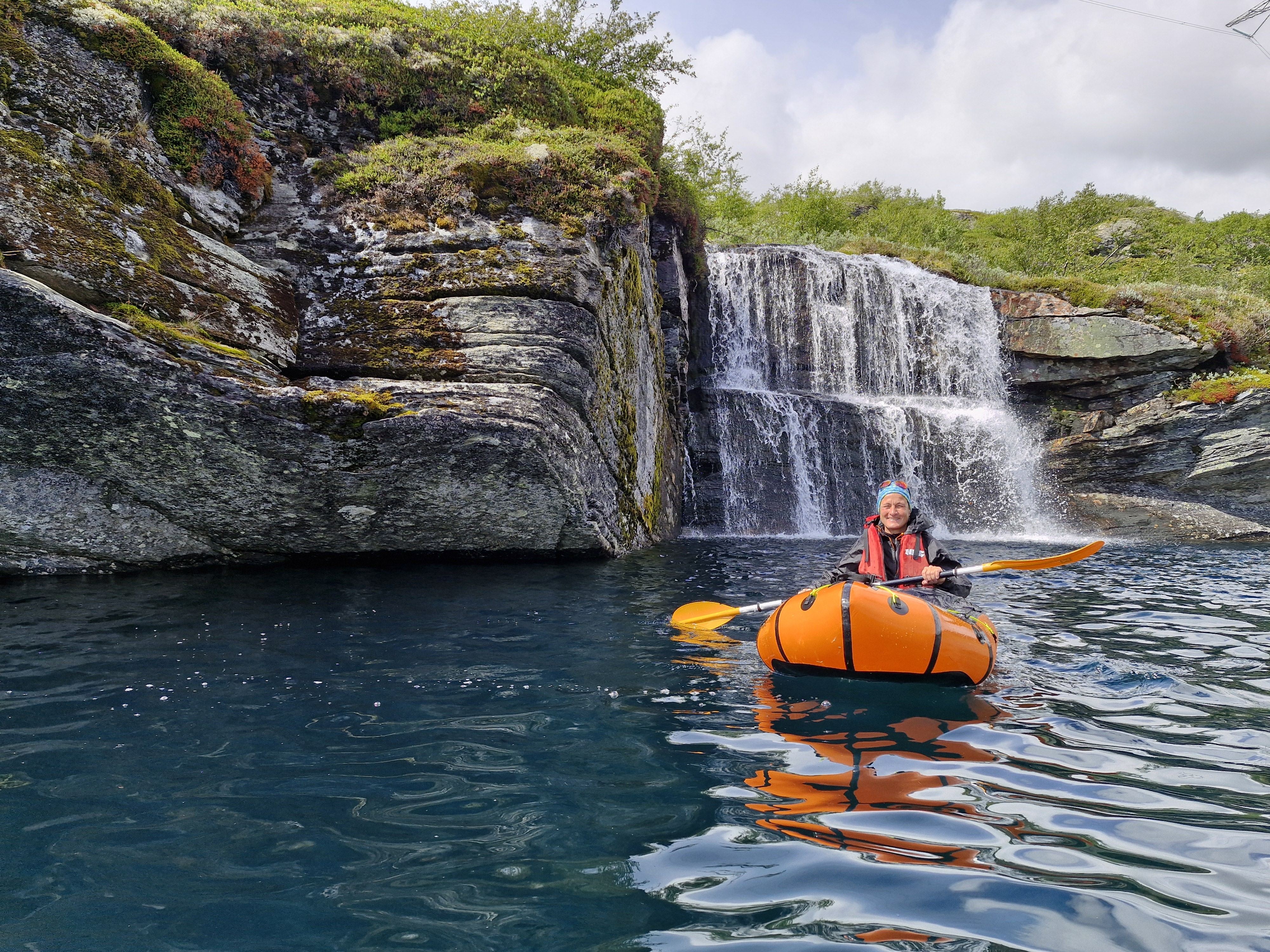 Ein person i ein packraft, på eit klart og blått fjellvatn. I bakgrunnen eit lite fossefall med dobbel dropp. 