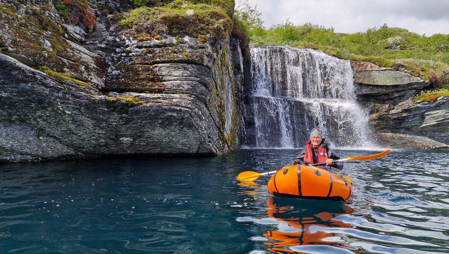 Ein person i ein packraft, på eit klart og blått fjellvatn. I bakgrunnen eit lite fossefall med dobbel dropp.