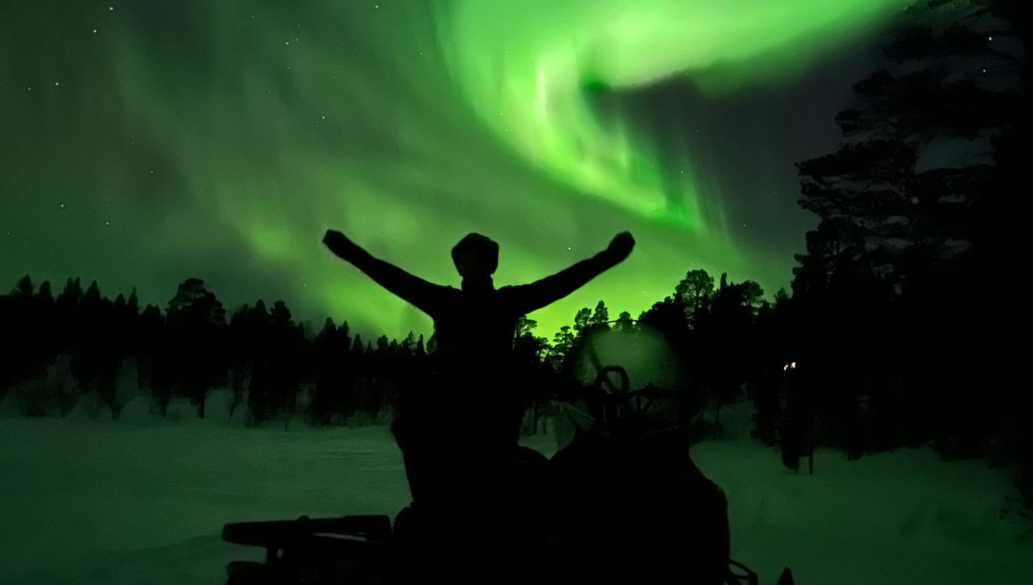 Experience the wonder of the northern lights dancing across the night sky. A silhouette of a person celebrates this breathtaking natural display while snowmobiles rest in the tranquil snow-covered landscape, creating a magical scene perfect for adventure