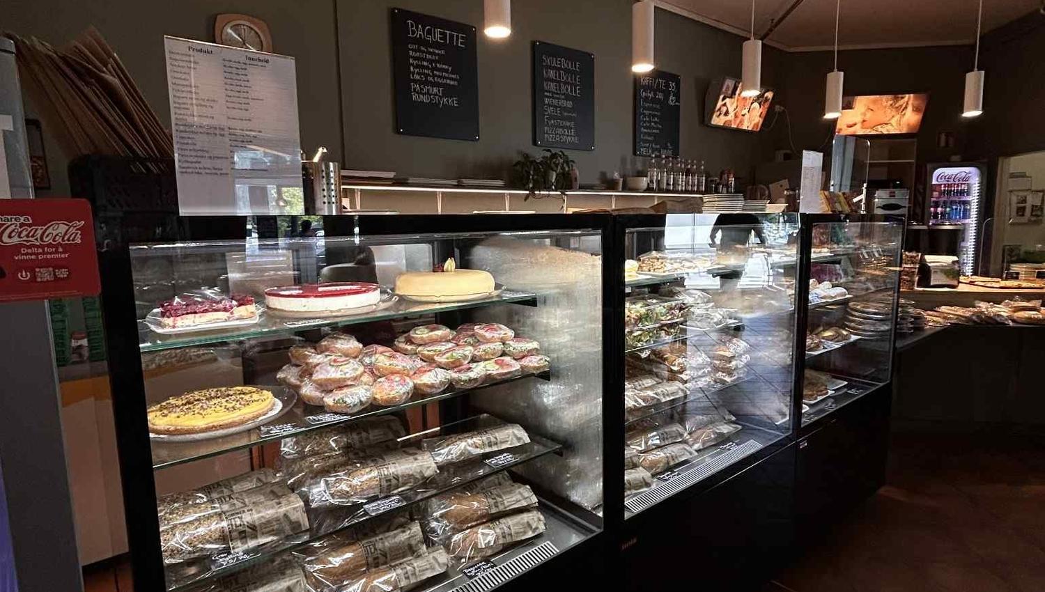 Interior of a bakery or cafe with glass display cases filled with pastries and cakes, and menu boards on the wall.
