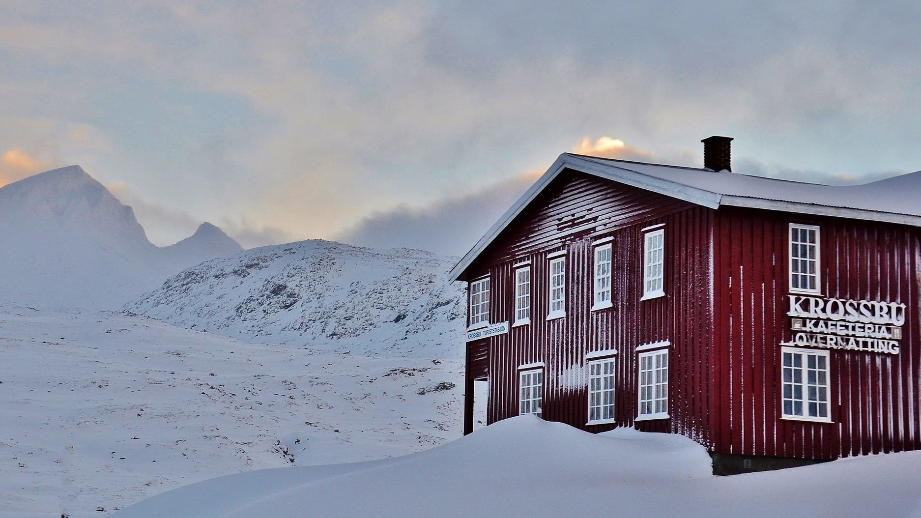 Red wooden lodge with white-trim windows beside deep snowdrifts, set against snowy mountains under a cloudy sky..