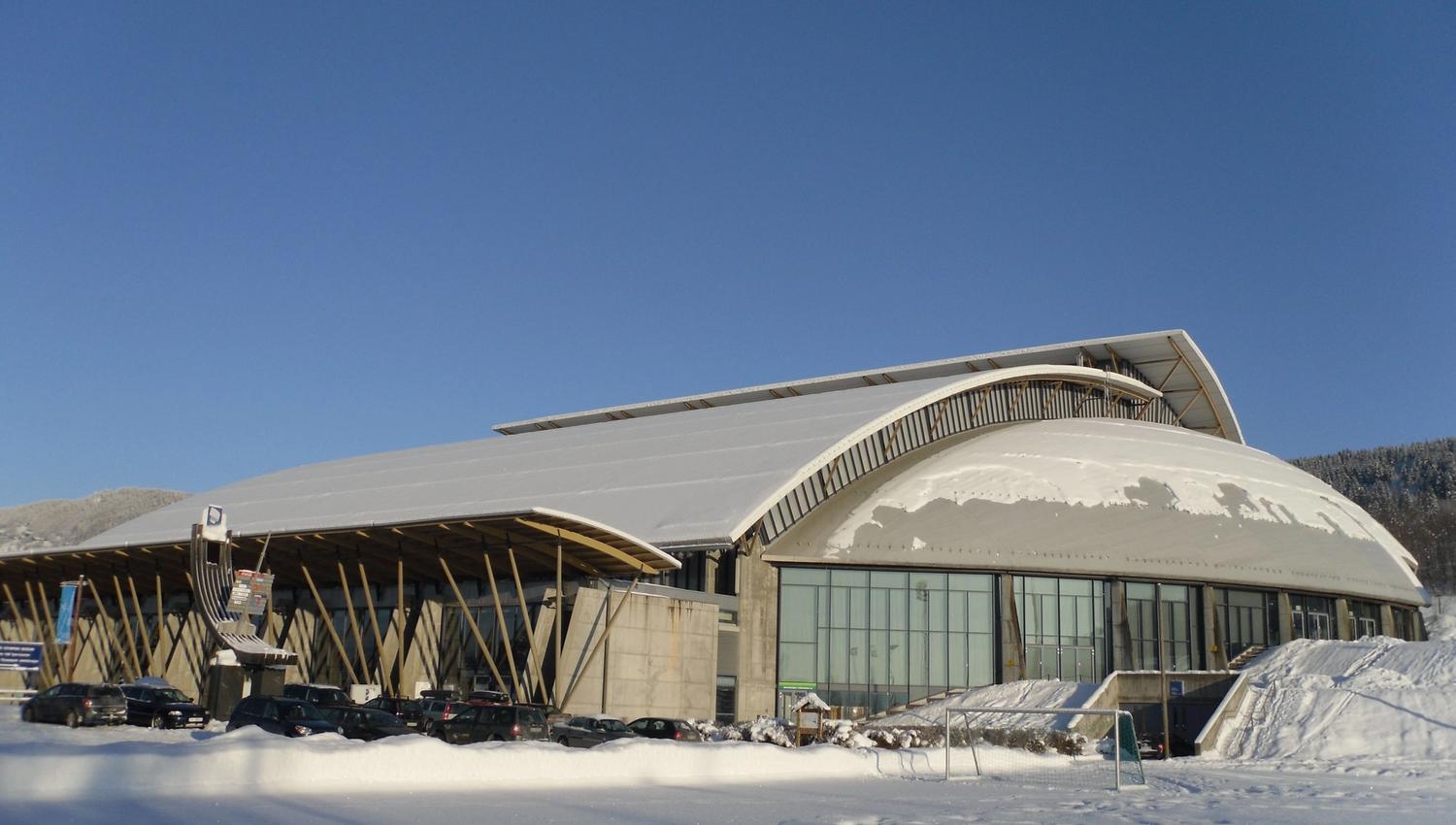 Stor moderne idrettshall med buet tak og snø foran, parkerte biler og klar blå himmel i vinterlys..