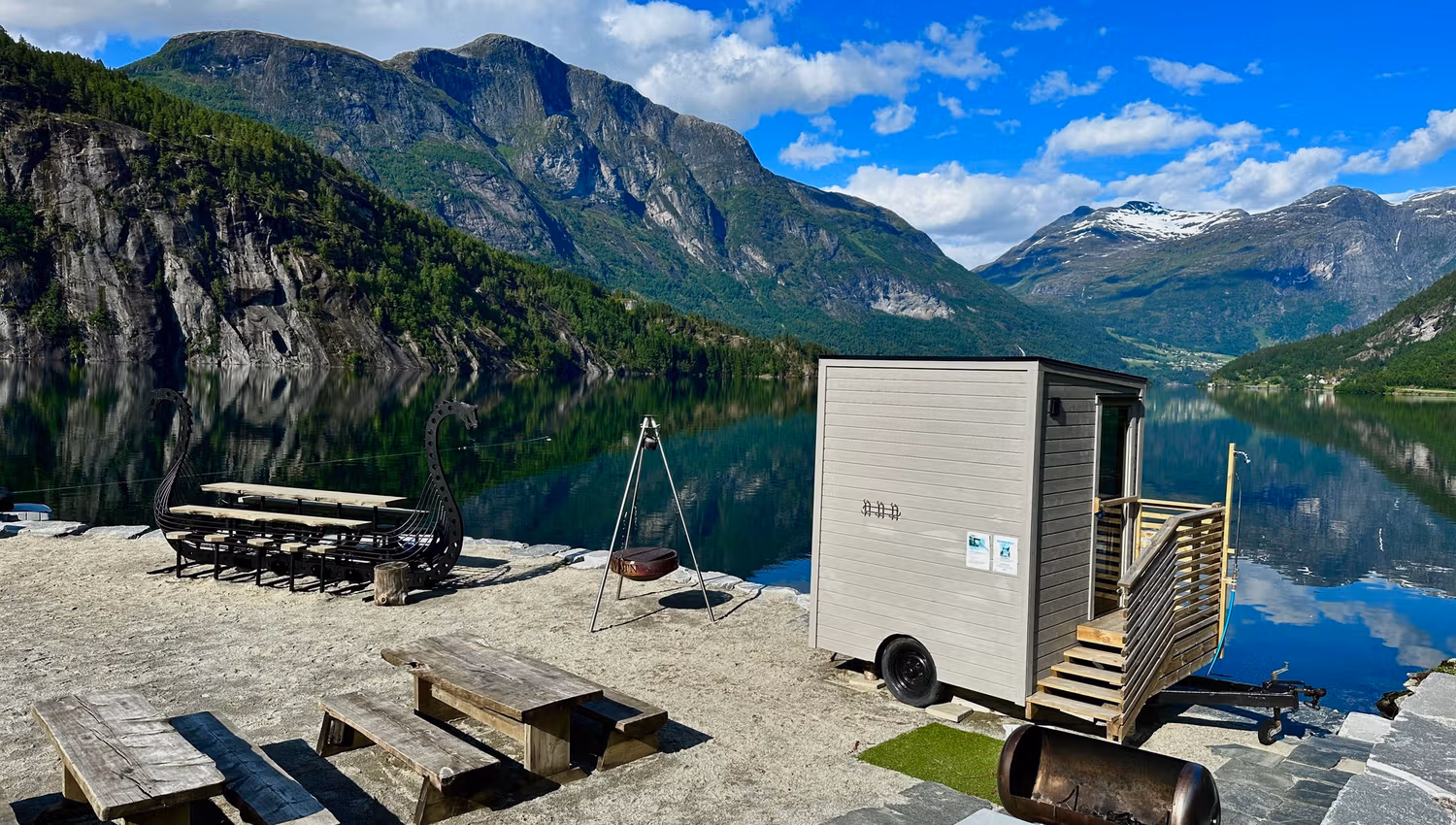 Small sauna cabin by a calm lake, with picnic tables, fire pit, and steep green mountains under blue sky..