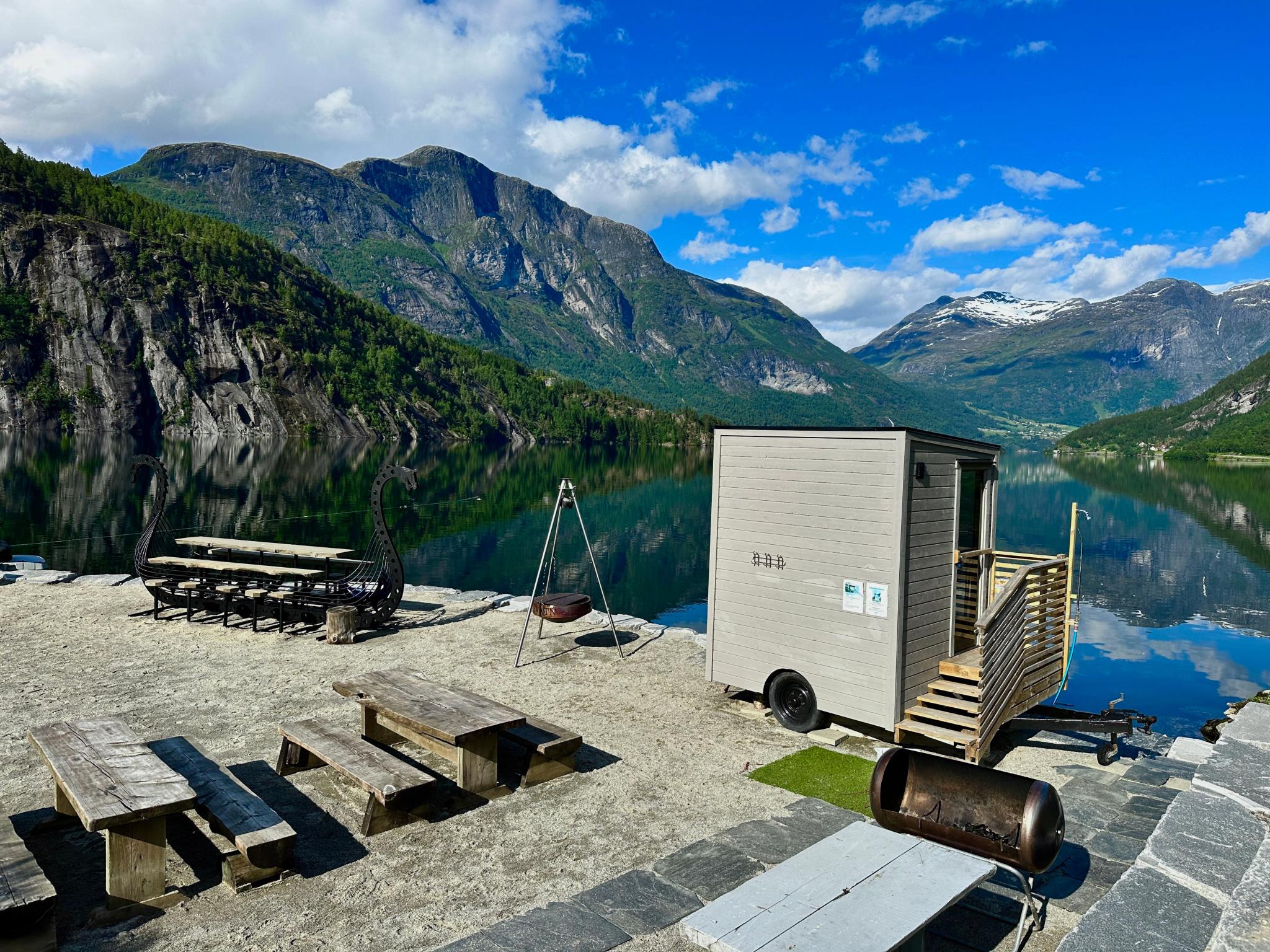 Small sauna cabin by a calm lake, with picnic tables, fire pit, and steep green mountains under blue sky..
