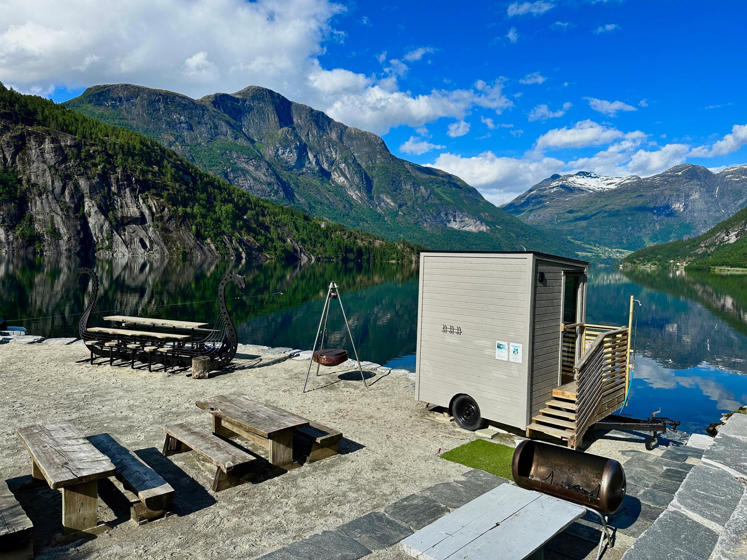 Small sauna cabin by a calm lake, with picnic tables, fire pit, and steep green mountains under blue sky..